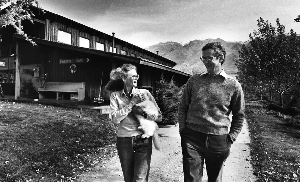 A black and white photo of a man and woman walking with a cat, with a building and mountains in the background.