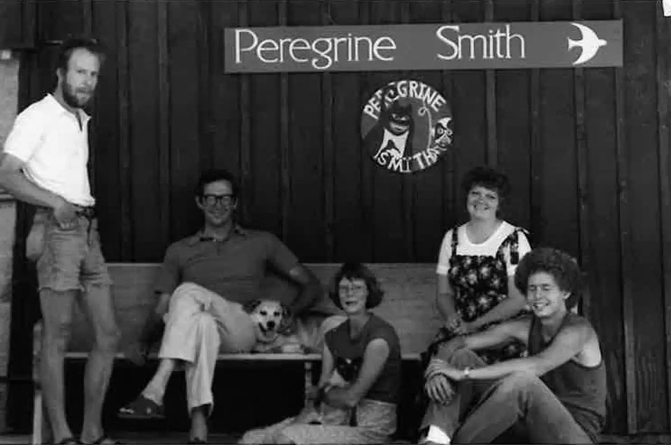 A group of people and dogs sitting and standing in front of a wooden wall with a sign.