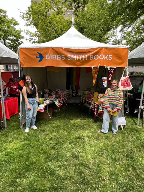 A white tent with an orange banner for ’GIBBS SMITH BOOKS’ at an outdoor market stall with two women standing in front of tables laden with books and merchandise.